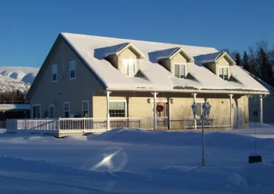 Two-story snow-covered house with dormer windows, wrap-around porch, and mountain backdrop in winter.