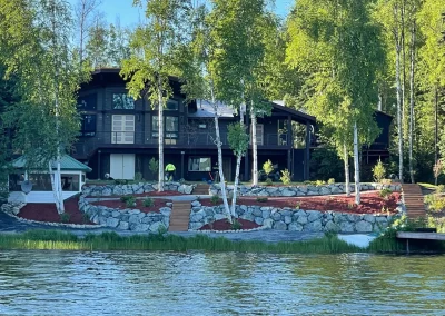 Modern lakefront home viewed from the water, featuring stone terraced landscaping and birch trees.